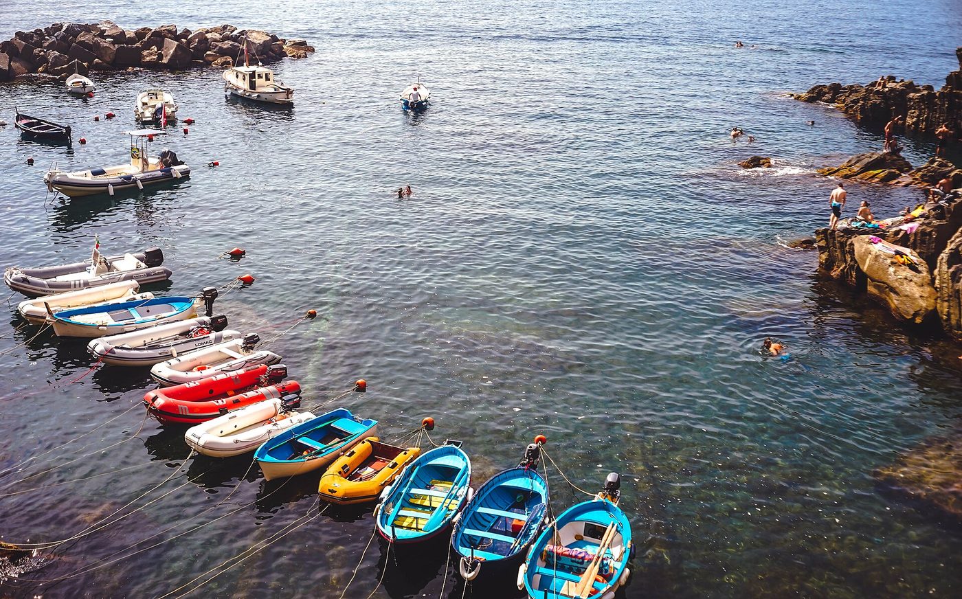 Small boats moored in a Cinque Terre harbour with swimmers on the rocks alongside